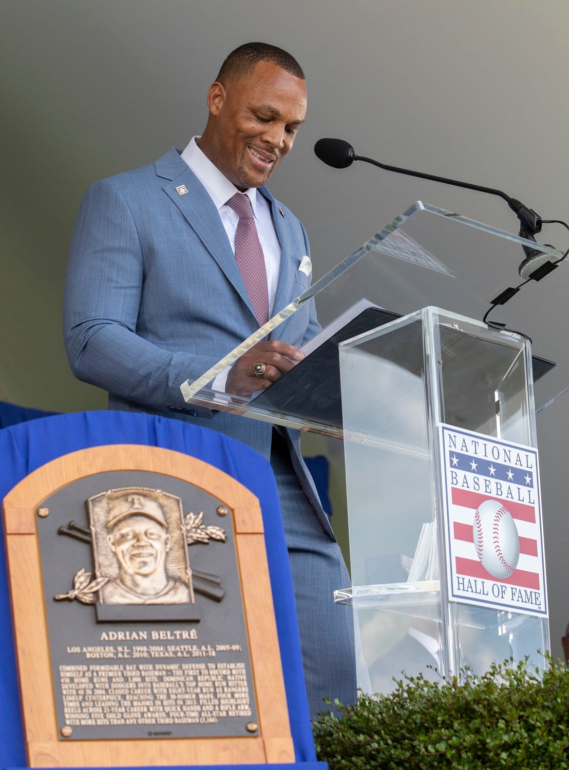 Adrian Beltre giving his HOF induction speech at a clear lexan podium, his HOF plaque is visible in the lower left of the photo